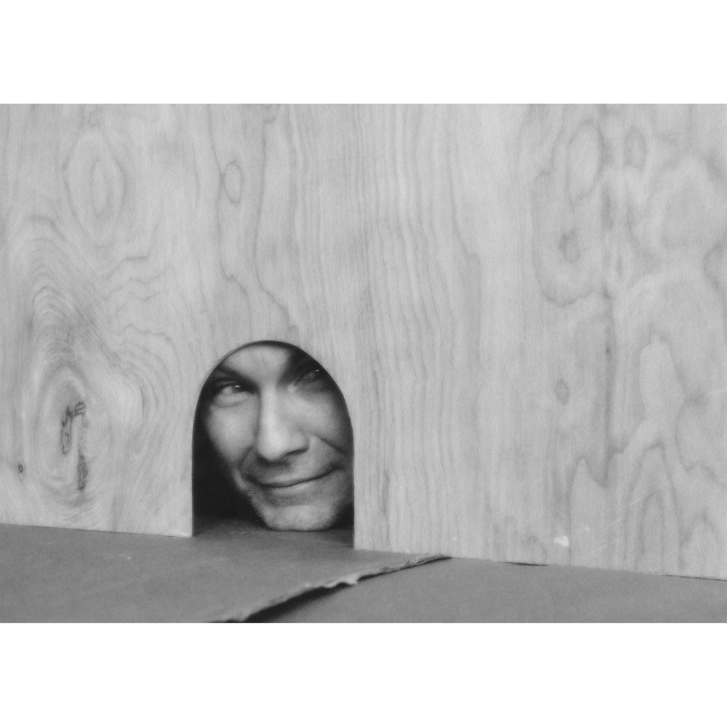 Black and white portrait of an adult male smiling and peering through an arched cutout in a wooden panel, medium shot.