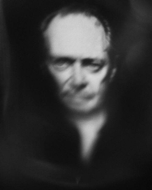 Black and white portrait of an adult man with fair skin and short hair staring directly at the camera in a blurred photo studio setting.