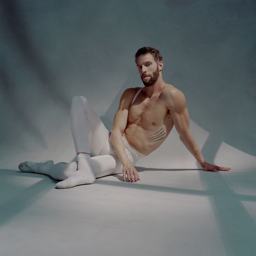 Athletic adult male in white leggings and bare torso poses on a seamless backdrop with studio lighting and shadows in a studio shot.