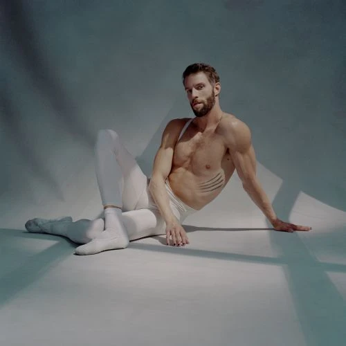 Athletic adult male in white leggings and bare torso poses on a seamless backdrop with studio lighting and shadows in a studio shot.