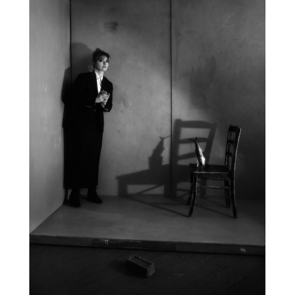 Black and white studio shot of a young adult woman in a dark suit leaning against a wall with chair and brick.
