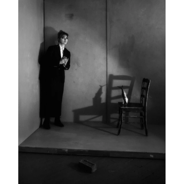 Black and white studio shot of a young adult woman in a dark suit leaning against a wall with chair and brick.