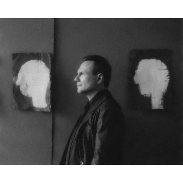 Monochrome portrait of an adult man in a dark coat, standing in front of wall art with head profile images in a minimalist style.