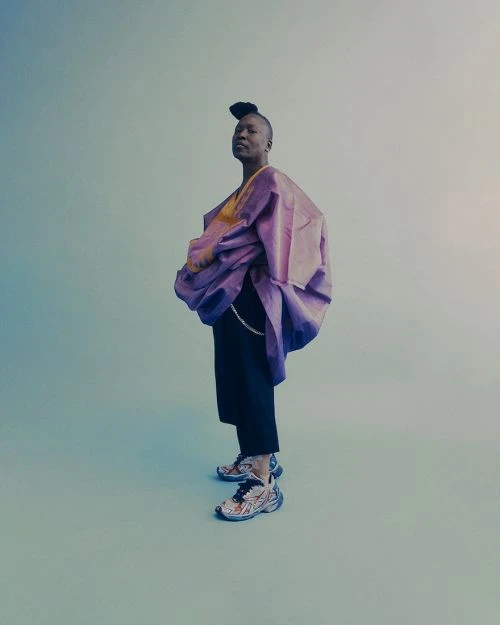 Person in a unique lavender-colored garment with culottes and colorful sneakers, posing in a studio against a gradient background.