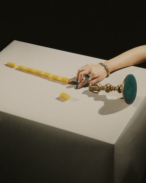 Overhead studio shot of a hand with turquoise jewelry resting on a white table next to an ornate brass candlestick and segmented candle.