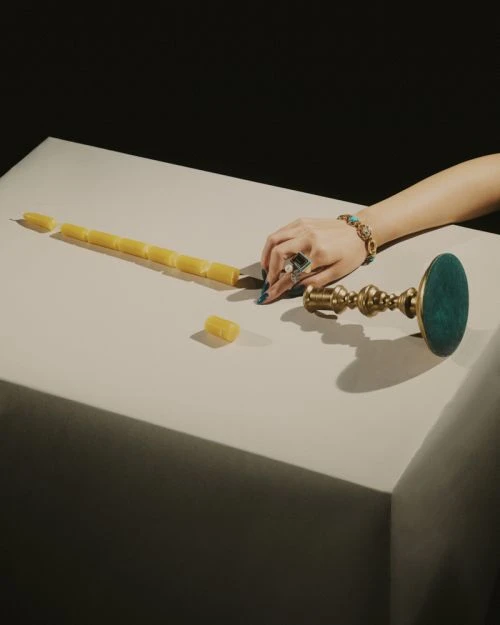 Overhead studio shot of a hand with turquoise jewelry resting on a white table next to an ornate brass candlestick and segmented candle.