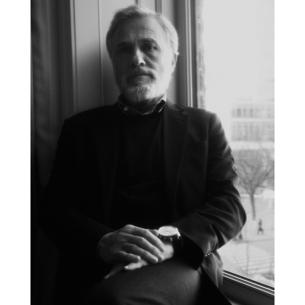 Portrait of an older man with white beard wearing a dark suit jacket seated near a window.