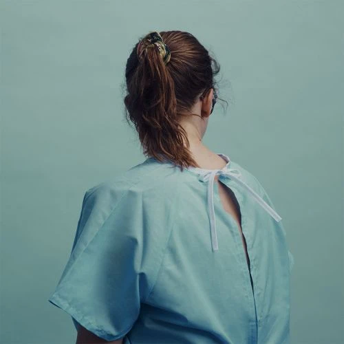 Back view of a woman with brown hair pulled back in a ponytail, wearing a blue hospital gown against a pale blue background.