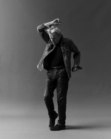 Monochrome portrait of an older man in a denim jacket and dark jeans dancing against a grey studio backdrop.