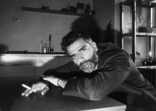 Portrait of Oscar Isaac leaning over a table in a domestic kitchen interior holding a cigarette, photographed in black and white by Erik Tanner
