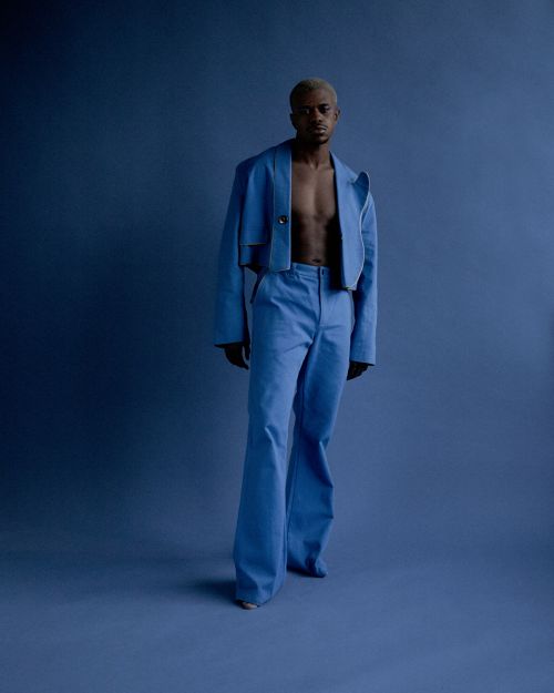 Man in a light blue suit stands against a blue background looking at the camera, creating a striking fashion editorial image.