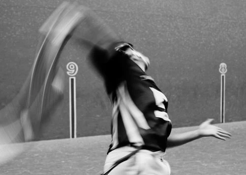 Action shot of an athlete swinging a cesta in a black and white indoor setting with number markings on the wall.