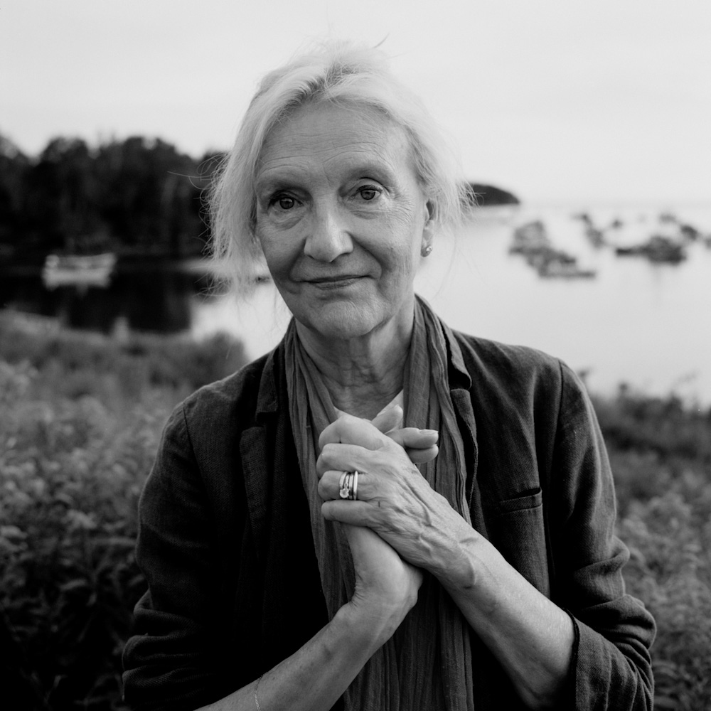 Portrait of an elderly woman with clasped hands wearing a jacket and scarf with an outdoor coastal background.