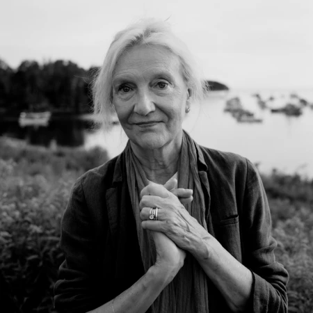 Portrait of an elderly woman with clasped hands wearing a jacket and scarf with an outdoor coastal background.