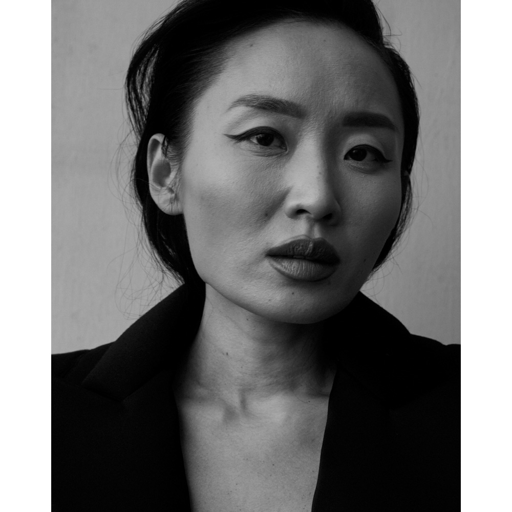 Monochrome portrait of an Asian woman with dark hair, bold makeup, and a black blazer, posing against a neutral wall background.