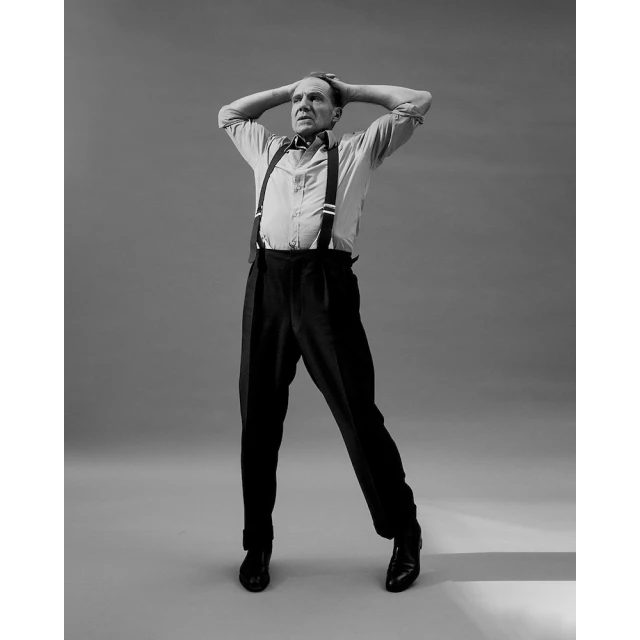 Full-length portrait of an adult man in a light-colored shirt, dark suspenders and pants, and black shoes, standing against a grey backdrop.