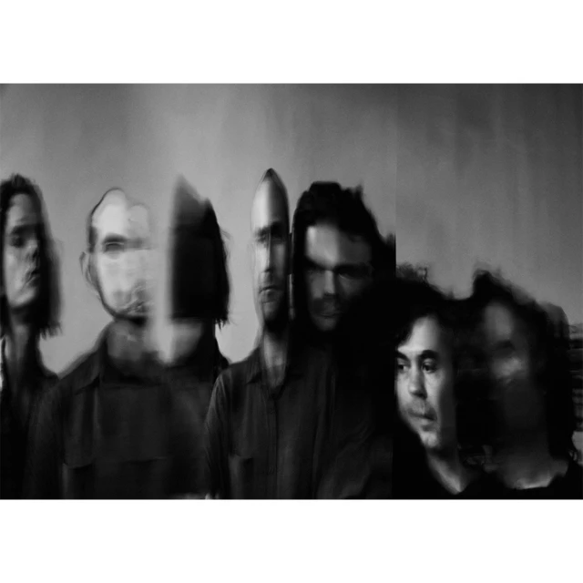 Black and white image of six blurred figures standing indoors against a light gray background, creating a soft, abstract portrait.