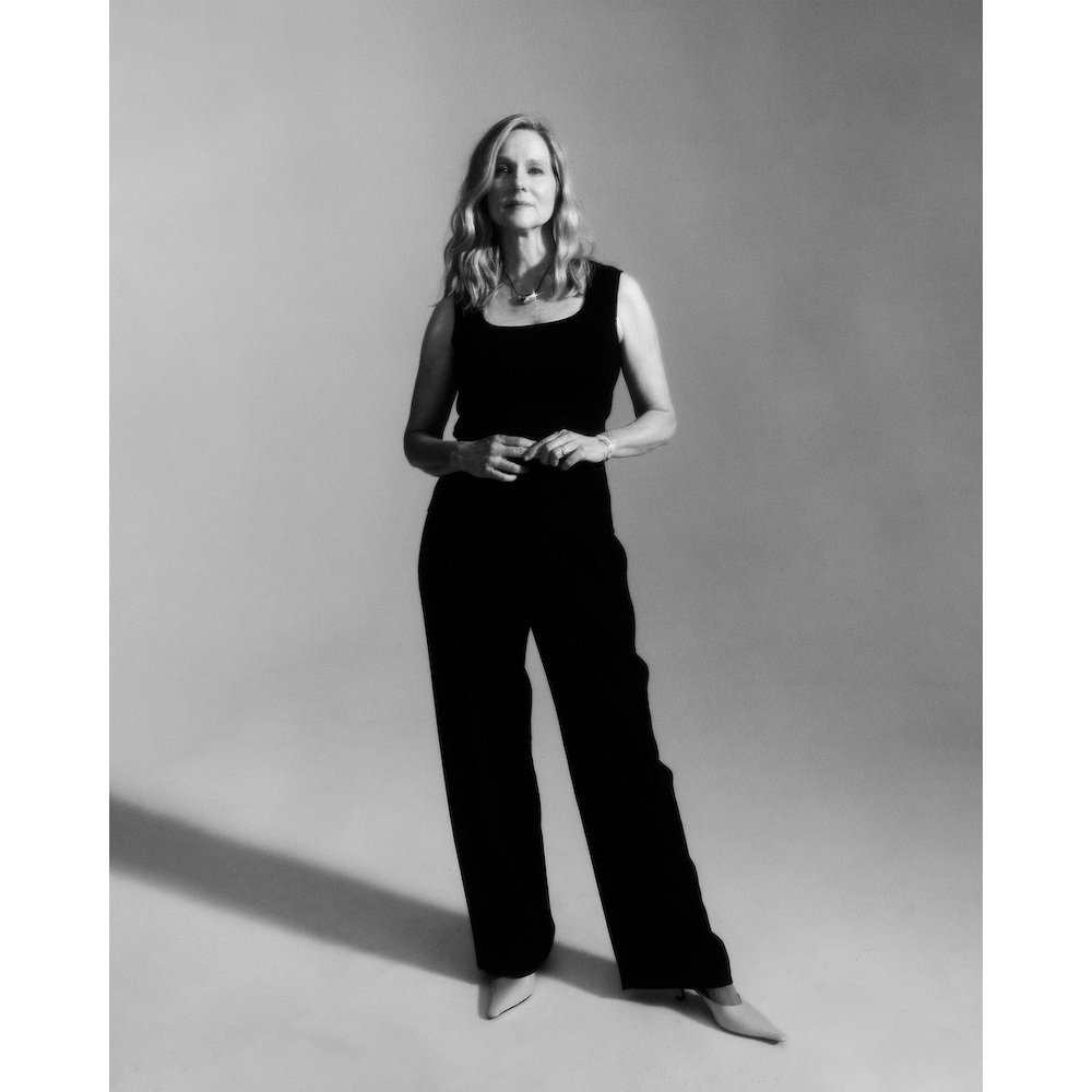 Elegant woman in a black top and wide-leg pants, accessorized with a necklace and heels, poses in a neutral studio setting.
