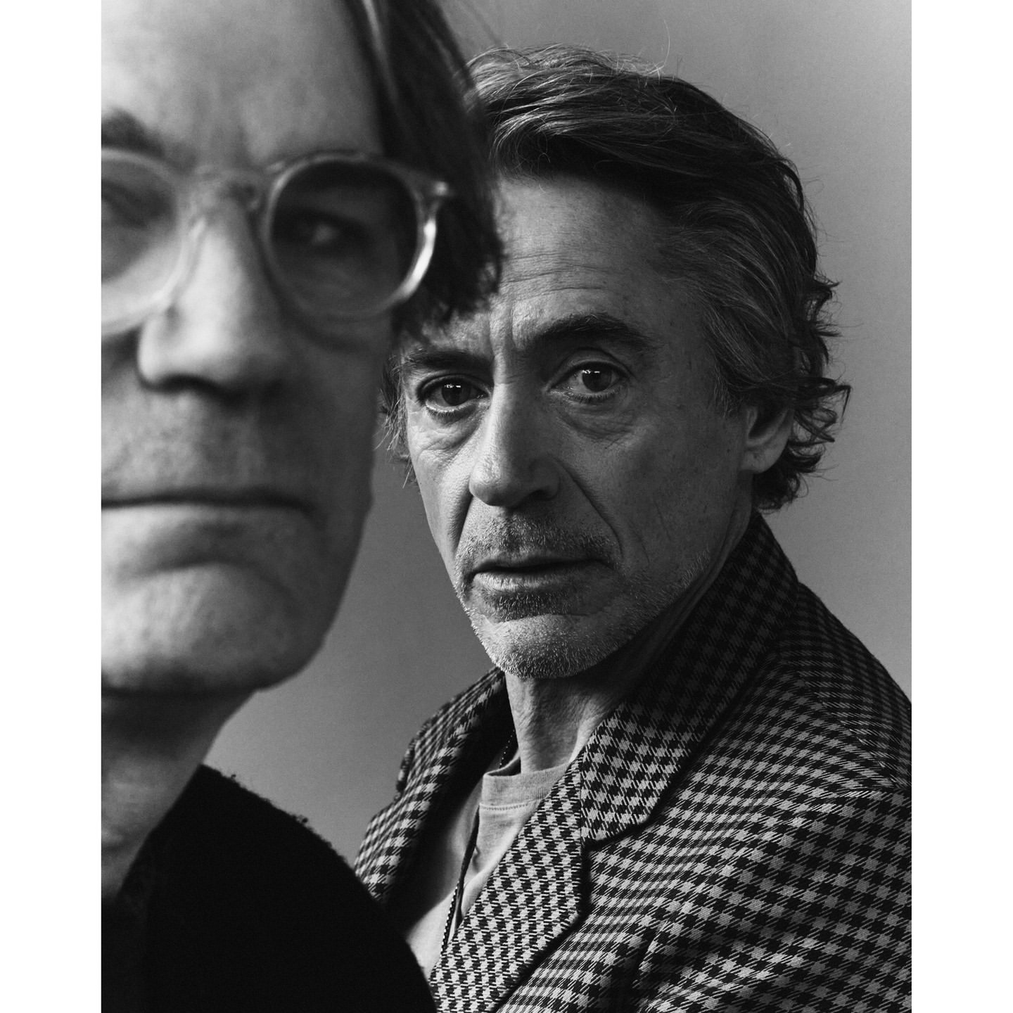 Monochrome portrait of two men, one in the foreground wearing round glasses and another in a patterned jacket behind him.