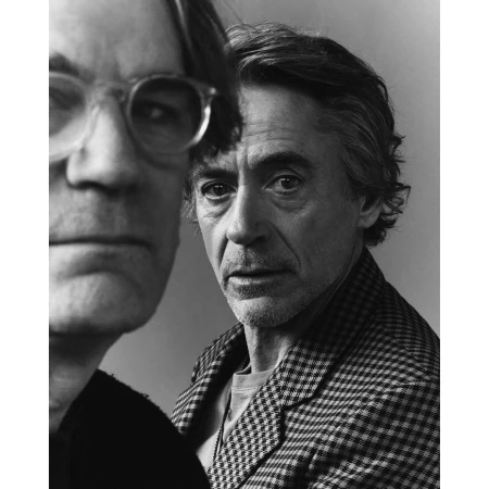 Monochrome portrait of two men, one in the foreground wearing round glasses and another in a patterned jacket behind him.