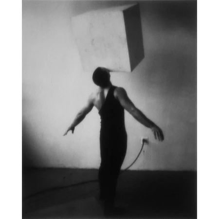 Monochrome photo of a man balancing a cube on his head, arms outstretched, against a plain wall with an electrical outlet and long cord.