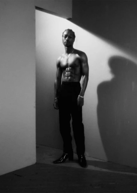 Striking black and white photo of a muscular, shirtless man in black pants and boots, standing in a corner of a white room with shadows.