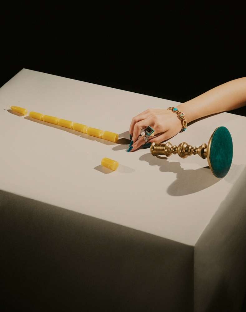 Overhead shot of a table with a woman's hand, jewelry, a segmented candle, and a brass candle holder against a dark background.