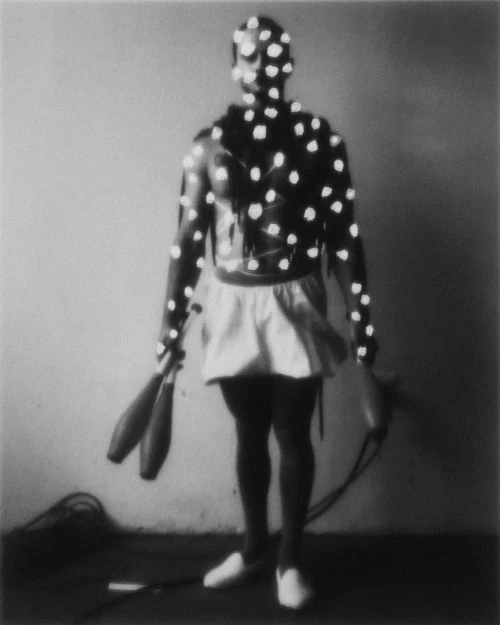 A black and white image of a muscular man with white dots on his body, holding juggling clubs in a dimly lit room. 