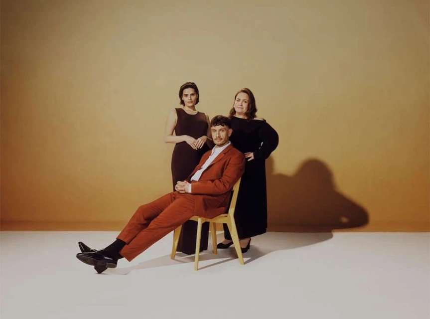 Group portrait of two women and one man, set against a tan backdrop in an indoor photo studio with soft, diffused lighting.