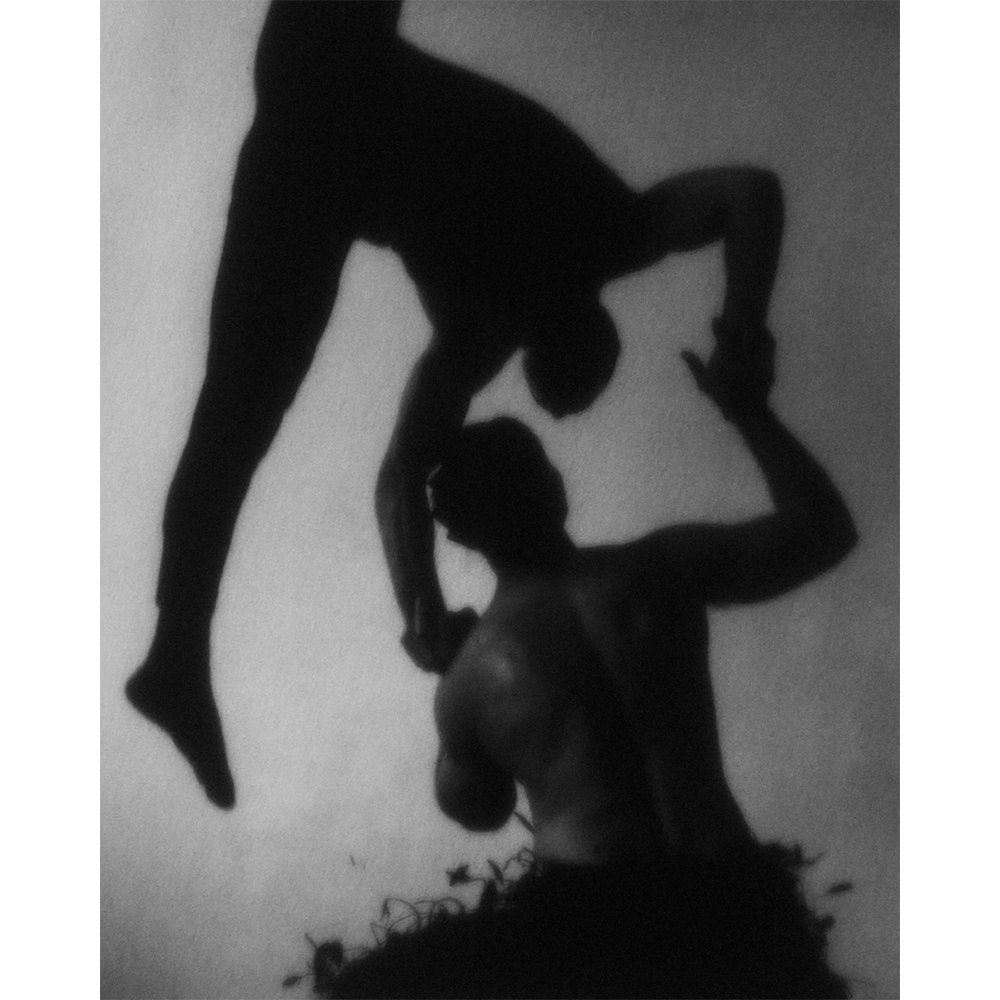 Monochrome photo of two silhouetted adult male figures in an acrobatic pose, one above the other, against a light background.