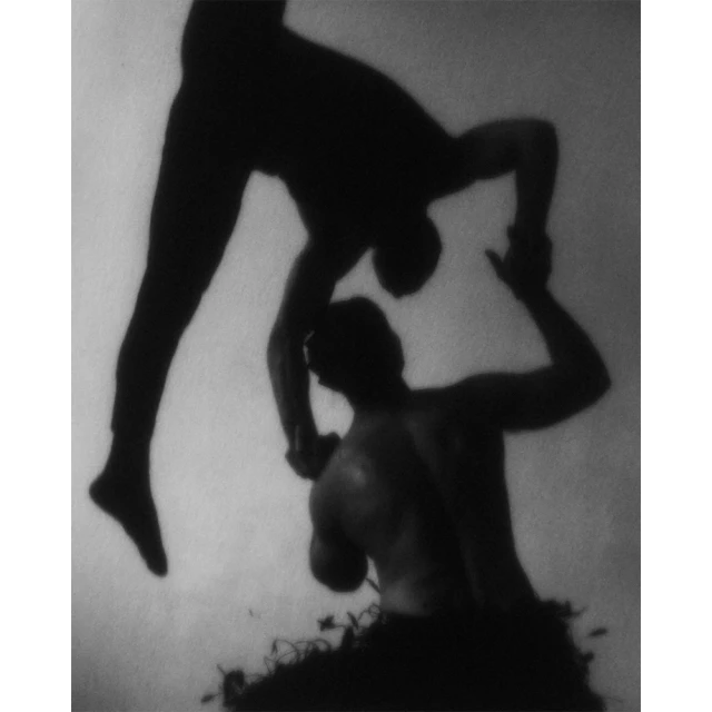 Monochrome photo of two silhouetted adult male figures in an acrobatic pose, one above the other, against a light background.