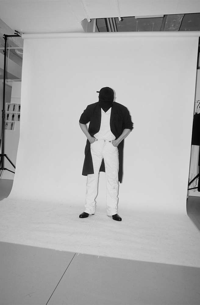 Monochrome studio photo of a person wearing a dark coat, white t-shirt, white jeans, and a cap against a white seamless backdrop.