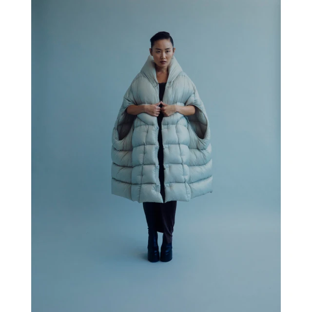 Full-length studio shot of a woman wearing a large quilted grey cape with black pants and black boots against a light grey backdrop.