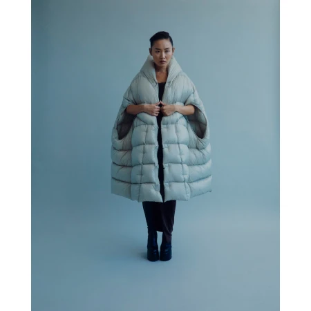 Full-length studio shot of a woman wearing a large quilted grey cape with black pants and black boots against a light grey backdrop.