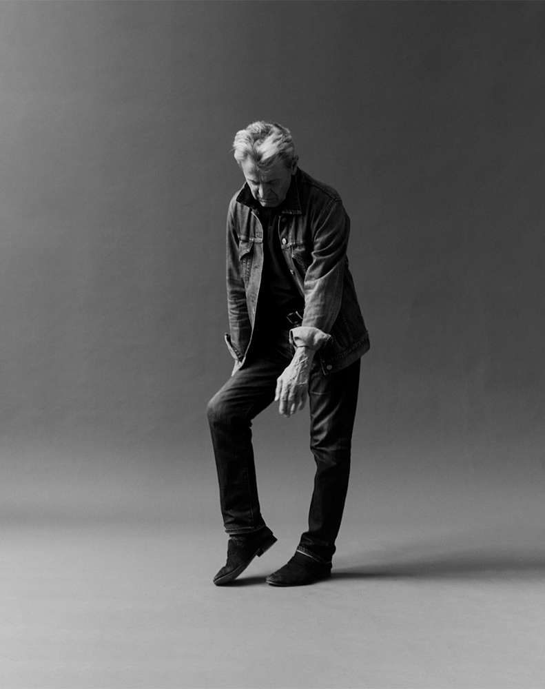 Black and white studio portrait of a mature adult male in a denim jacket and jeans standing with one leg resting, looking down.