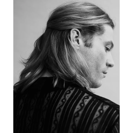 Monochrome portrait of a man with long hair in half-up style and patterned dark shirt against light background by Erik Tanner