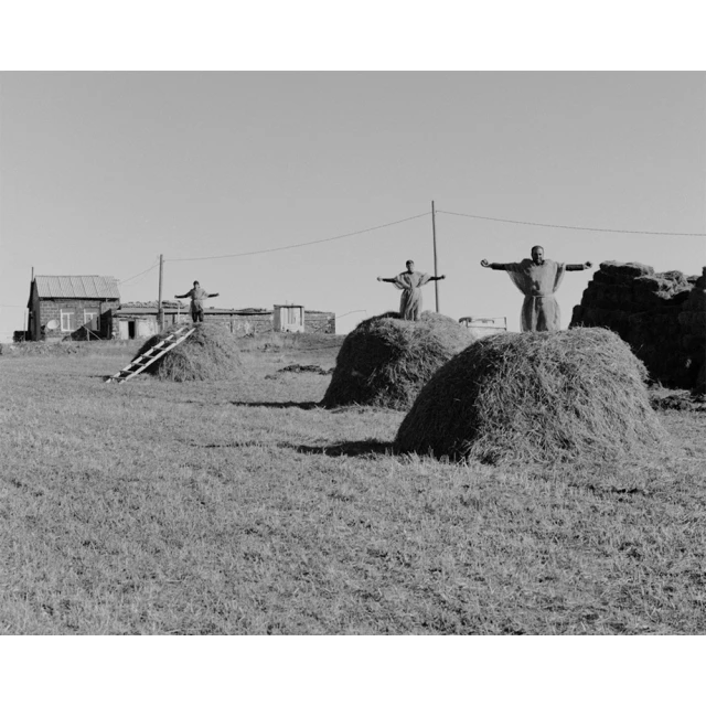 Black and white landscape featuring three people in robes standing on haystacks with arms outstretched, buildings in the background.
