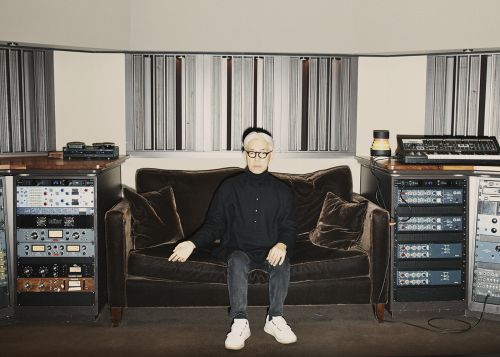 Mature adult male seated on a brown couch in a music studio surrounded by sound equipment and wearing glasses and a black shirt.