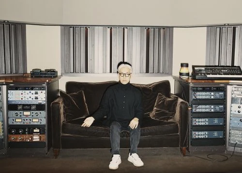 Mature adult male seated on a brown couch in a music studio surrounded by sound equipment and wearing glasses and a black shirt.