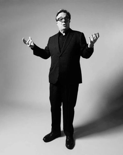 Full-length studio shot of a man in a pinstripe suit and clerical collar standing with open hands in front of a neutral background.