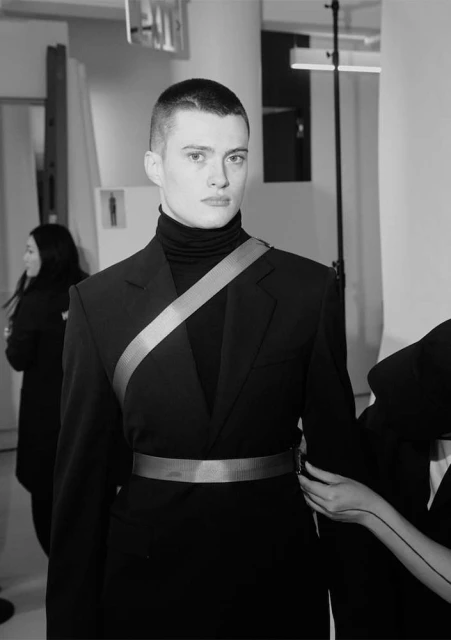 Monochrome studio shot of a young adult man in a dark suit with a gold belt and sash, posing with neutral expression, partial view of others.
