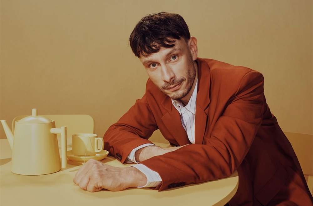 Portrait of an adult man wearing a red blazer seated at a yellow table with a tea set in a softly lit, neutral-toned indoor setting.