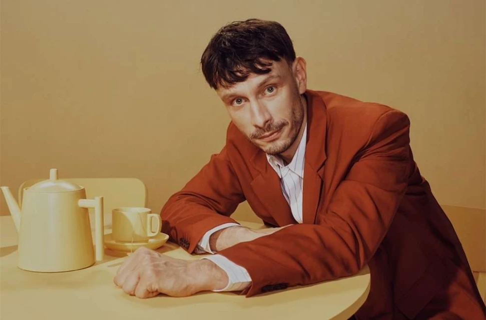 Portrait of an adult man wearing a red blazer seated at a yellow table with a tea set in a softly lit, neutral-toned indoor setting.