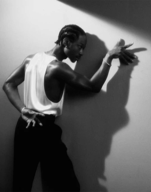 Black and white portrait of a Black male model leaning against a wall, wearing a tank top and trousers, posing in a studio with strong lighting.