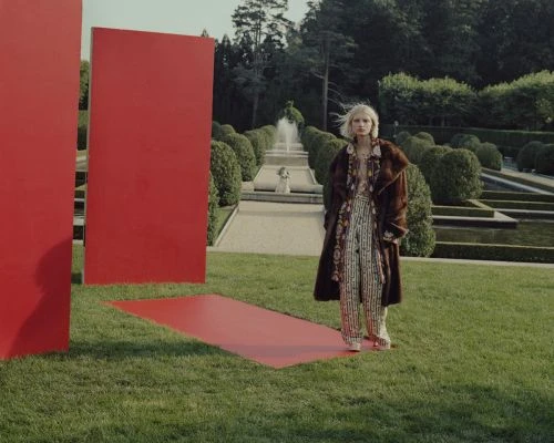 Fashion shot in a garden with a woman in a patterned outfit and fur coat standing near red rectangular panels and a fountain.