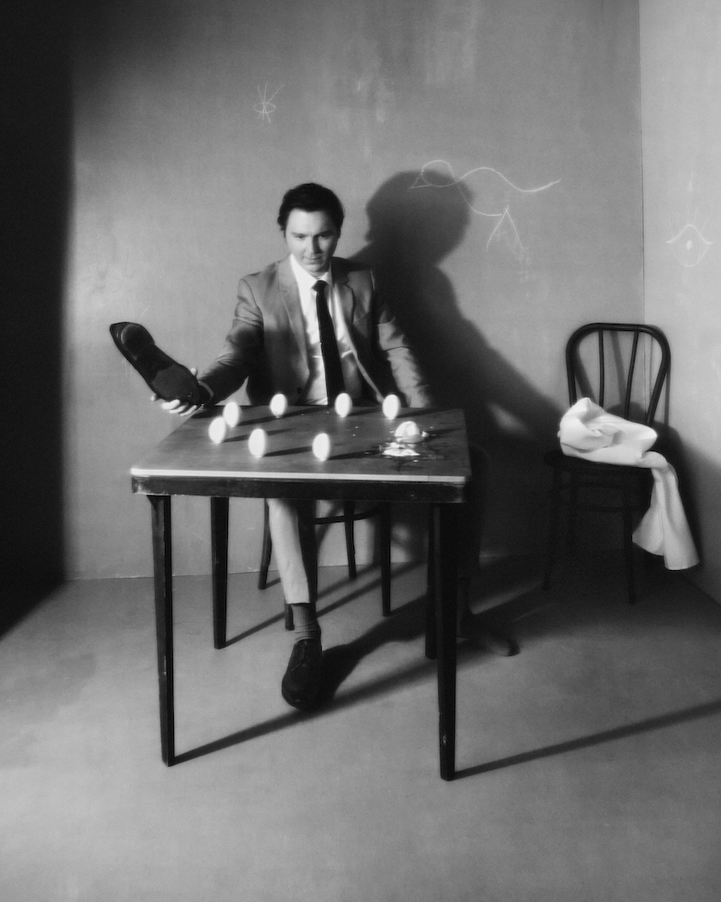 Black and white photo of a man at a table with eggs, holding a shoe. The scene is set indoors with simple lighting.