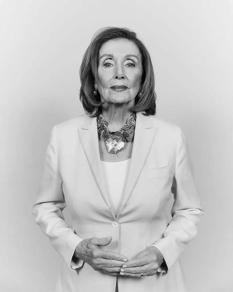 Portrait of Nancy Pelosi wearing a light blazer and patterned necklace against a plain studio background.