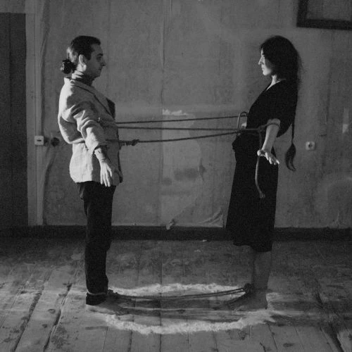 Black and white image of an adult man and woman facing each other, bound together with rope in a sparse interior setting with a painted backdrop.