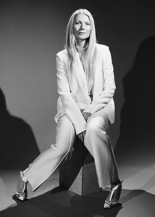 Blond woman in a white suit seated on a block in a photo studio. The photo is black and white and taken in a medium shot.