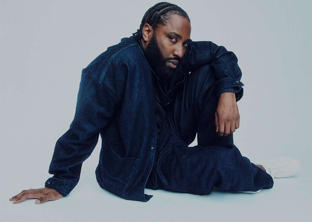 Portrait of a stylish Black male model with braided hair, posing in a denim jacket and pants on a light background in a studio setting.